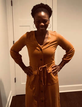 Picture African American with an Afro and big round earrings, in a cognac colored dress, standing against a white wall/door and hardwood floor in the background with both hands on waist line 